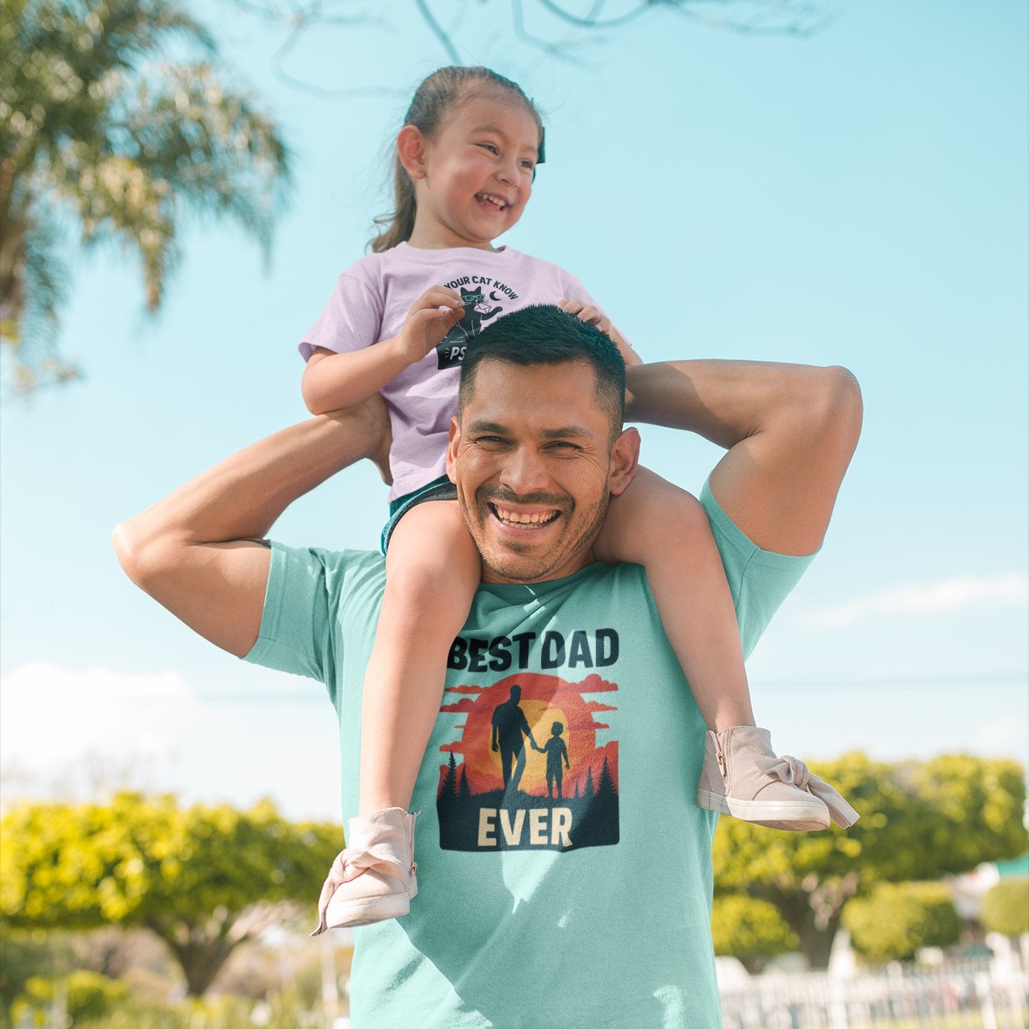 Dad wearing Best Dad Ever shirt with child on shoulders, smiling in sunny park setting