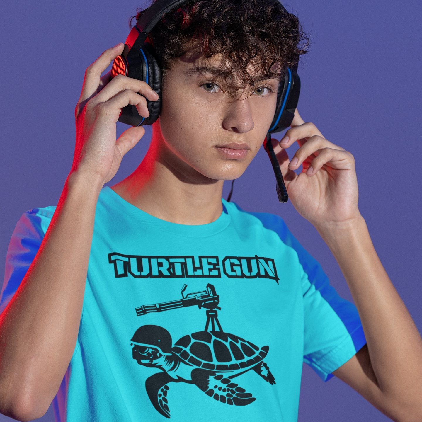 Young man wearing a bright blue t-shirt with turtle gun design, holding headphones and smiling.